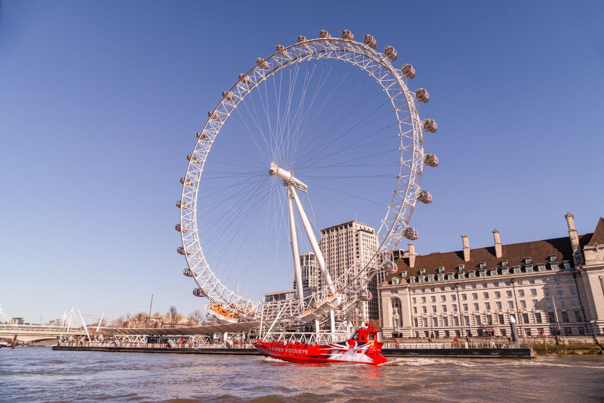 The London Eye in the sun, viewed from the water with a Thames Rockets speedboat passing by.